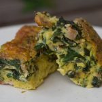 Two slices of a frittata are propped on a plate. The top is golden brown, while green leaves and yellow eggs interleave the inside.